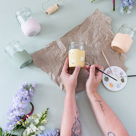 Upcycling-Idee: DIY-Vasen aus Einmachgläsern gestalten - Step 2: Einmachglas mit Blüten bemalen Zwei Hände bemalen ein gelb angemaltes Einmachglas mit Hilfe eines Pinsels mit weißen Blüten.
