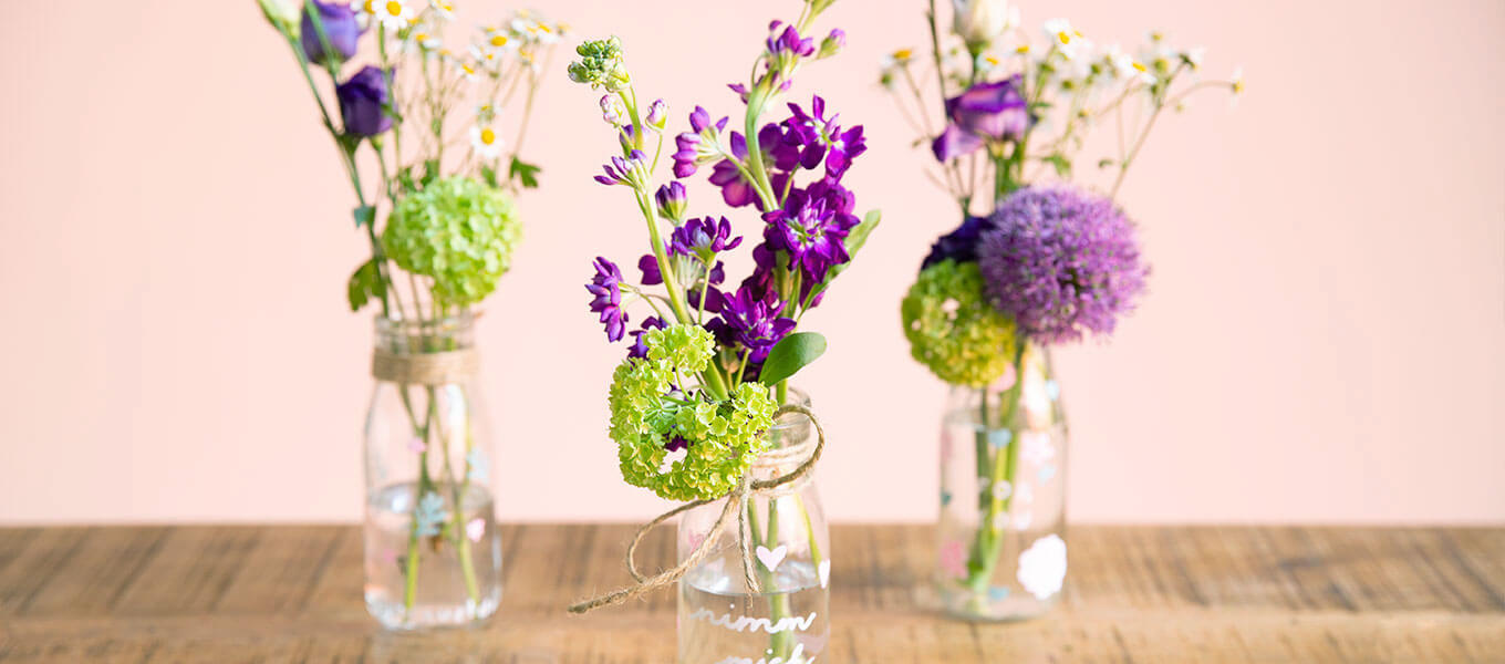 Drei Einmachgläser mit Blumen arrangiert stehen auf einem Tisch. Auf den Einmachgläsern stehen verschiedene Botschaften, welche mit Kreidefarbe aufgebracht worden sind. 
