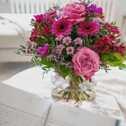 ein Buch ragt ins Bild und im Hintergrund steht ein pinker Blumenstrauß in einer Vase