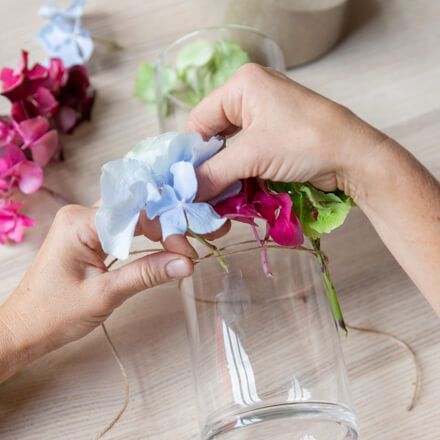 Deko mit Hortensien: Windlicht dekorieren - Step 2: Hortensienblüten nach und nach am Glas befestigen Zwei Hände befestigen einen kleinen Teil einer Hortensienblüte zwischen zwei Juteschnüren.