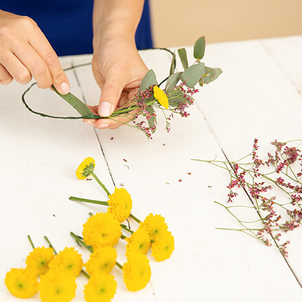 DIY: Haarkranz selbst basteln - Step 3: Blumen am Kranz befestigen Zwei Hände wickeln mit Hilfe von Floristentape Santini, Eukalyptus und Strandflieder an einen Haarkranz aus Draht.