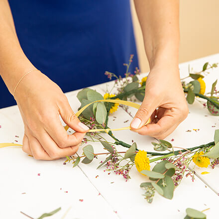 Zwei Hände befestigen an einen mit Blumen geschmückten Haarkranz ein dünnes Band in Gelb.