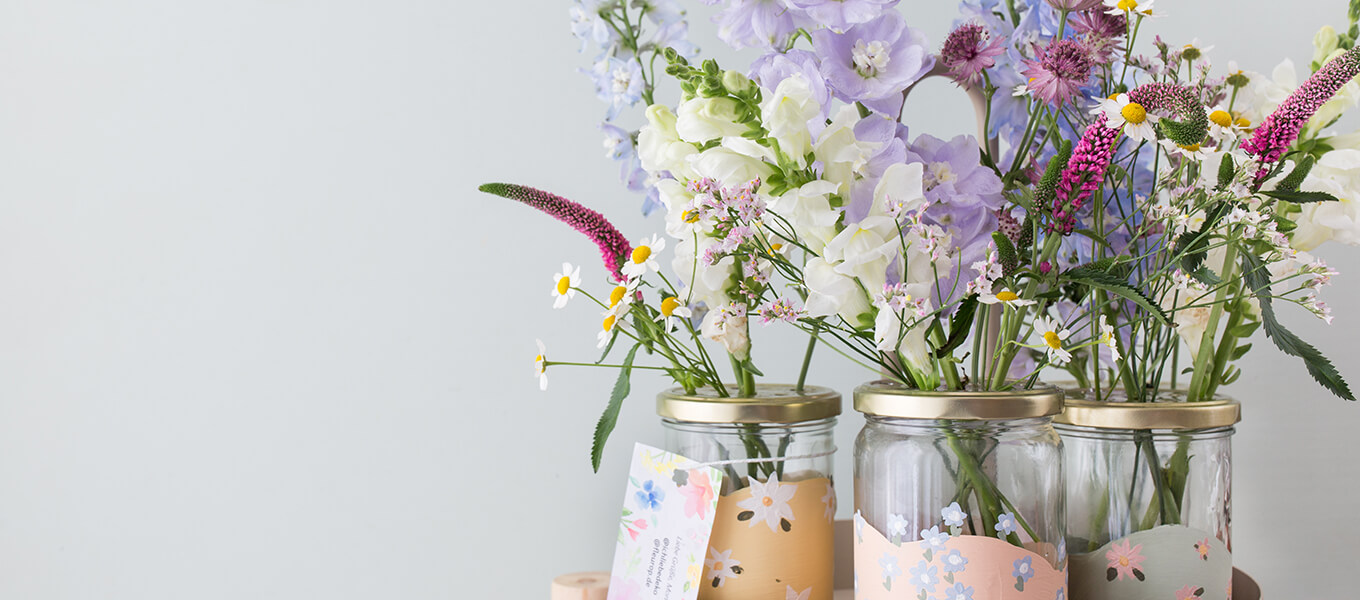 Upcycling-Idee: DIY-Vasen aus Einmachgläsern gestalten Mehrere mit Kreidefarbe angemalten Einmachgläser stehen auf einem Tablett und sind mit vielen schönen Blüten befüllt. Die Einmachgläser sind zudem dezent mit Blüten bemalt. An dem einen Einmachglas ist ein Zettel mit einer Grußnachricht befestigt.