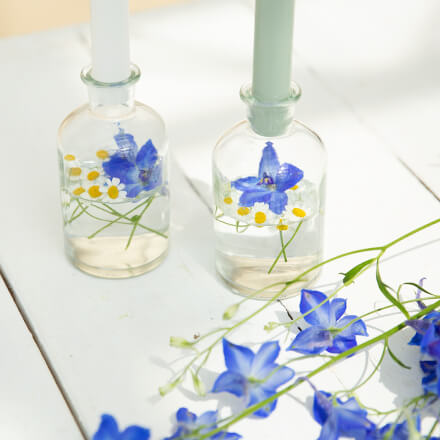 Einfache Tischdeko für den Frühsommer - DIY Nahaufnahme von zwei Gingläsern, die mit etwas Wasser, sowie Kamille- und Ritterspornblüten befüllt sind. Auf den Ginflaschen steckt eine weiße und eine mintfarbene Kerze. Neben den zwei Gingläsern liegt etwas Rittersporn zur Dekoration.