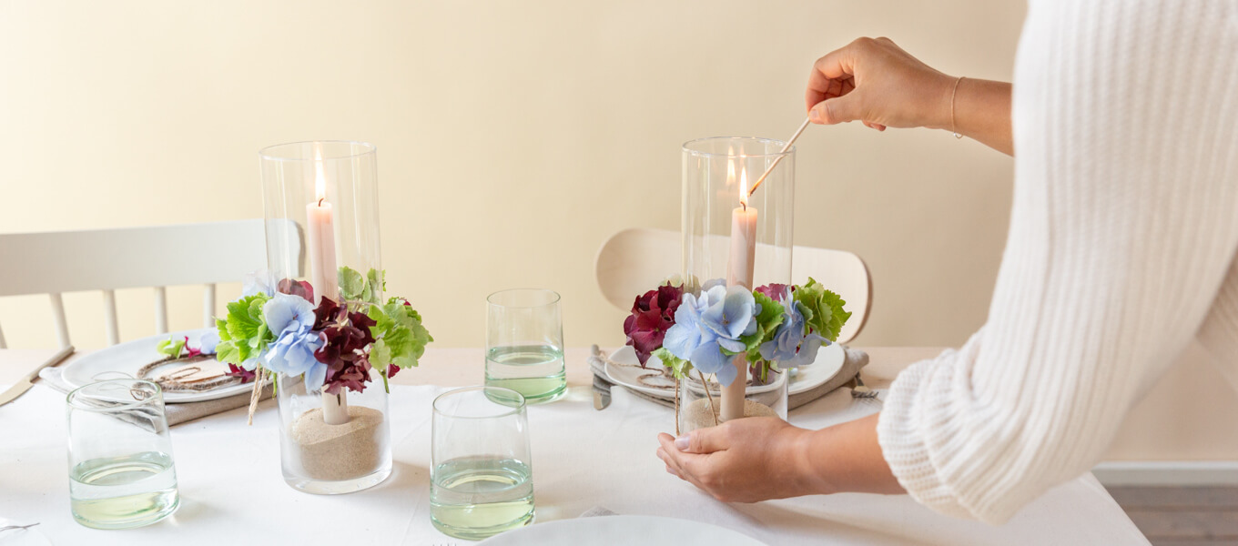 Tischsituation, wobei eine Person Windlicht anzündet, welches am äußeren Rand des Glases mit Hortensien dekoriert ist.