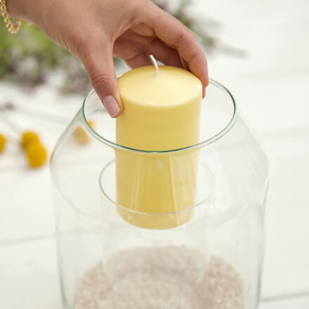 Dekoidee: Windlicht für den Sommertisch - Step 2: Kerze einsetzen Eine Hand dekoriert ein sich in einem Windlicht befindlichen Zylinder mit einer gelben Kerze.