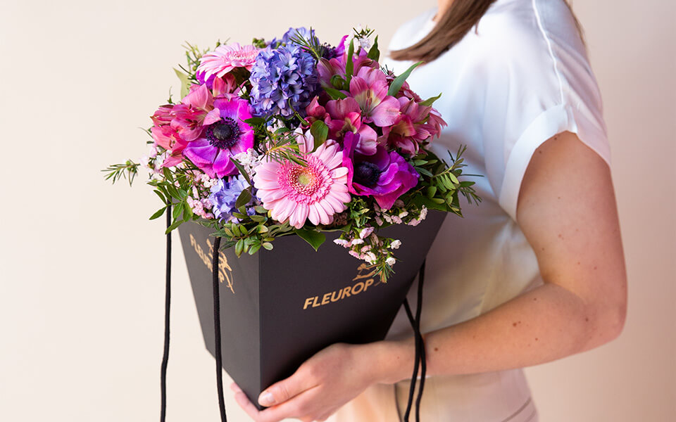 Eine Frau hält eine Flower-Bag in der Hand. In der Flower-Bag ist ein lila, pink, rosafarbener Blumenstrauß drinnen.