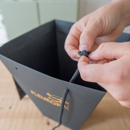 Flower-Bag-Upcycling-Idee zur Geschenkbox: Step 1 Die schwarzen Henkel einer Flowerbag werden von zwei Händen entfernt.