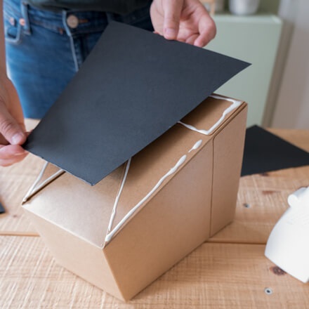 Flower-Bag-Upcycling-Idee zur Vase: Step 3 Eine auf der Seite liegende Flower-Bag wird mit einer zugeschnittenen Pappe in Schwarz beklebt auf einer Seite, wo zuvor Kleber aufgetragen wurde.