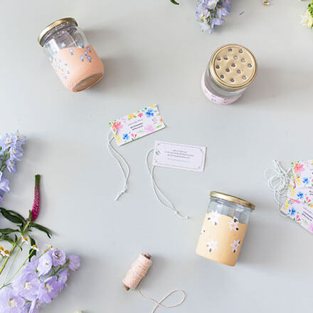 Upcycling-Idee: DIY-Vasen aus Einmachgläsern gestalten - Step 3: Deckel in Steckhilfe umwandeln Zwei Lonely-Bouquet-Day Anhänger sind zu sehen. Außerdem zwei Einmachgläser die mit dezenter Kreidefarbe angemalt sind und auf denen kleine Blüten aufgemalt worden. Zusätzlich wird ein Einmachglas von oben gezeigt, welches im Deckel mehrere Löcher hat.