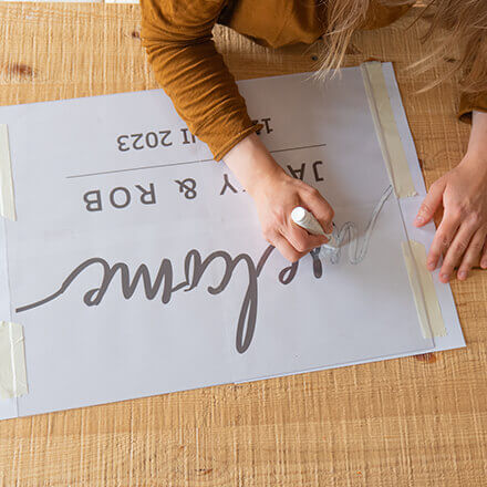 Willkommensschild Hochzeit: Step 2 Die Schrift eines Willkommensschilds wird von einer Vorlage auf die Scheibe eines großen Bilderrahmens abgepaust