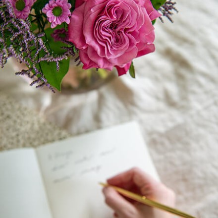 eine Hand schreibt in ein Notizbuch und in der Ecke steht ein Blumenstrauß