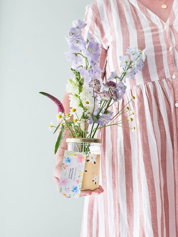 Eine Person hält mit einer Hand ein Einmachglas, welches mit schönen Blüten gefüllt ist. Das Einmachglas wurde mit gelber Kreidefarbe und dezenten weißen Blüten bemalt. An dem Einmachglas hängt eine kleine Karte mit einer Botschaft.