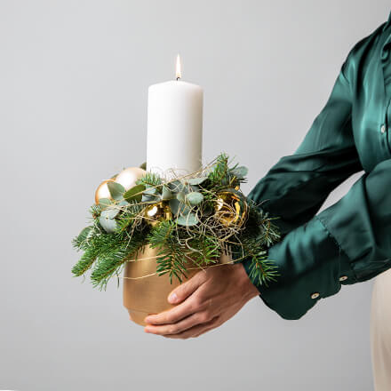 Modernes Adventsgesteck mit Stumpenkerze Modernes Adventsgesteck mit weißer Stumpenkerze, Eukalyptus, Tanne, goldenen und beigen Kugeln und einem goldenen Topf. Das Gesteck wird von einer Person in einem edlen dunkelgrünen Oberteil gehalten.