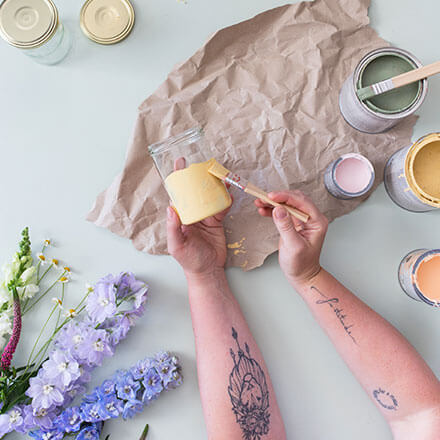 Upcycling-Idee: DIY-Vasen aus Einmachgläsern gestalten - Step 1: Einmachglas mit Kreidefarbe bemalen Zwei Hände bemalen ein Einmachglas mit Hilfe eines Pinsels mit gelber Kreidefarbe an.