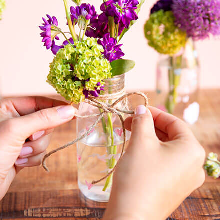 Zwei Hände binden eine Schleife mit Hilfe eine Juteschnur um ein Einmachglas, welches mit Blüten gefüllt ist. Auf dem Einmachglas steht "Nimm mich mit!" mit Kreidefarbe geschrieben.