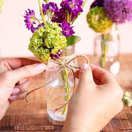 Blumen-Überraschung mit liebevoller Botschaft - Step 3: Glas verzieren und Anhänger befestigen Zwei Hände binden eine Schleife mit Hilfe eine Juteschnur um ein Einmachglas, welches mit Blüten gefüllt ist. Auf dem Einmachglas steht "Nimm mich mit!" mit Kreidefarbe geschrieben.