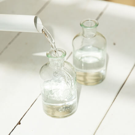 In zwei kleine Ginflaschen wird etwas Wasser mit Hilfe einer Gießkanne gefüllt.