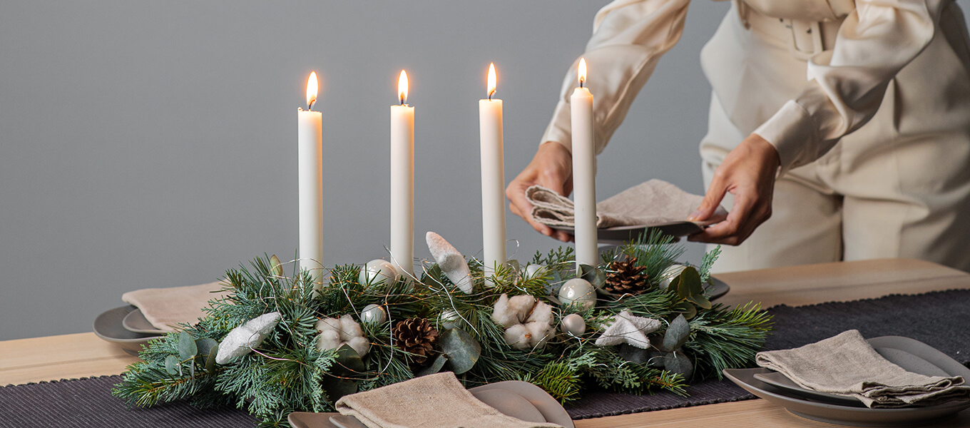 Deko für den Advent Modernes, längliches Adventsgesteck in grün weiß mit Stabkerzen. Eine Person dahinter dekoriert den Tisch weiter.