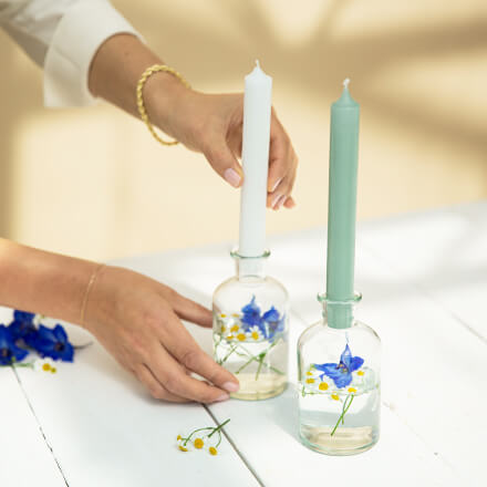 Einfache Tischdeko für den Frühsommer - Step 5: Kerzen hinzufügen Eine weiße Kerze wird auf ein Ginglas gesteckt, welches mit etwas Wasser, Kamille- und Ritterspornblüten gefüllt ist. Daneben steht eine weitere Kerze mit einem Ginglas. Diese Kerze ist in einem Min Ton.