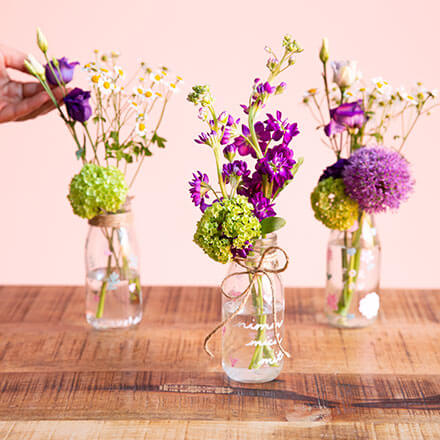 Blumen-Überraschung mit liebevoller Botschaft - fertiges DIY Auf einem Tisch stehen drei Einmachgläser mit unterschiedlichen Botschaften, welche mit Kreidefarbe darauf geschrieben worden sind. In den Einmachgläsern befinden sich Blumen. Eine Hand arrangiert weitere Blumen in eins der hinteren Einmachgläser.
