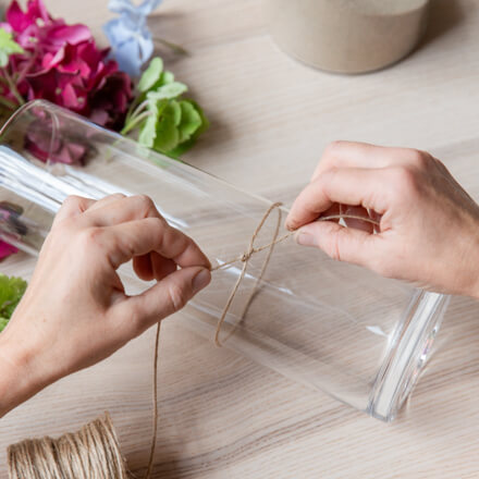 Deko mit Hortensien: Windlicht dekorieren: Step 1: Juteschnur am Glas befestigen Zwei Hände befestigen eine Juteschnur mit einem Knoten um ein Zylinderglasgefäß.