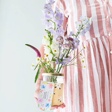 Upcycling-Idee: DIY-Vasen aus Einmachgläsern gestalten - Grußnachricht befestigen Eine Person hält mit einer Hand ein Einmachglas, welches mit schönen Blüten gefüllt ist. Das Einmachglas wurde mit gelber Kreidefarbe und dezenten weißen Blüten bemalt. An dem Einmachglas hängt eine kleine Karte mit einer Botschaft.