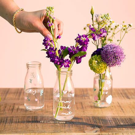 Blumen-Überraschung mit liebevoller Botschaft - Step 2: Blumen im Glas arrangieren Auf einem Tisch stehen drei Einmachgläser mit drei unterschiedlich aufgemalten Botschaften. Eine Hand arrangiert einzelne Blüten in das mittlere Glas, während das rechte Glas schon vollständig mit Blumen gefüllt ist. Das linke Glas ist noch leer.