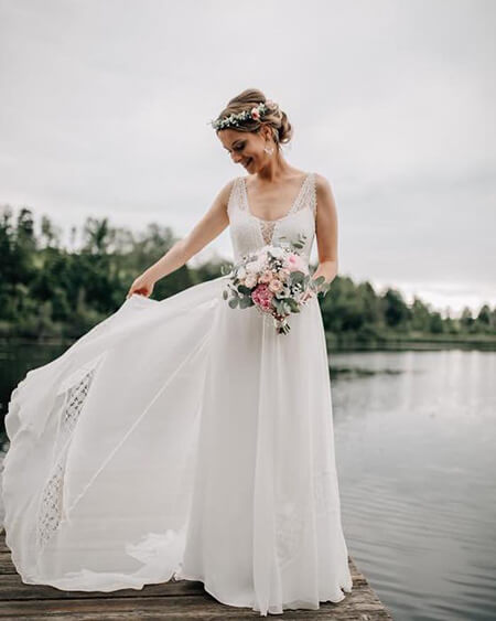 Braut steht auf einem Steg am See und hält einen traditionellen Brautstrauß