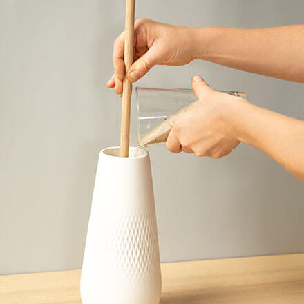 Ein langer Holzstanb wird in eine Vase gestellt. Anschließend wird Sand in die Vase befüllt, damit der Holzstab senkrecht stehen bleibt.