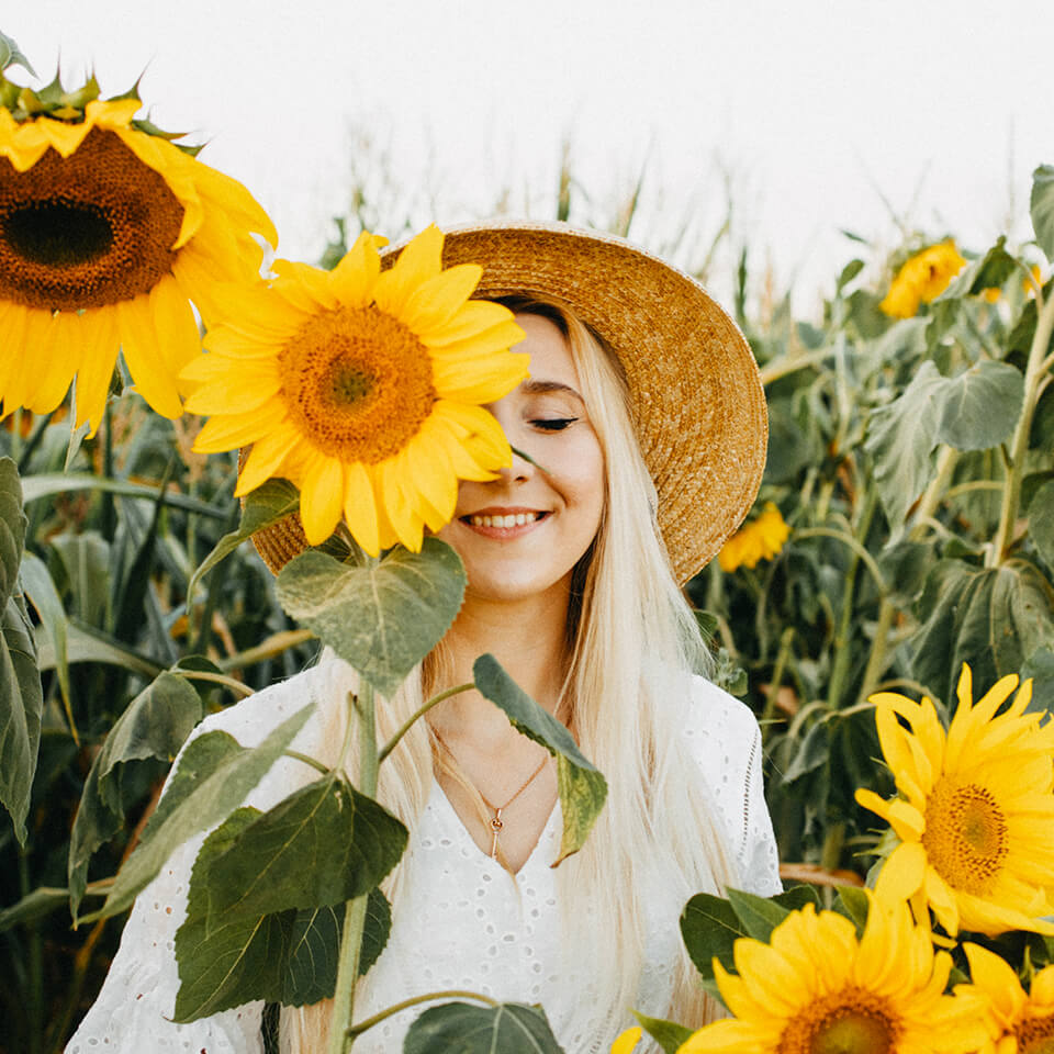 Eine Frau steht im Sonnenblumenfeld mit dem halben Gesicht hinter einer Sonnenblume und lächelt